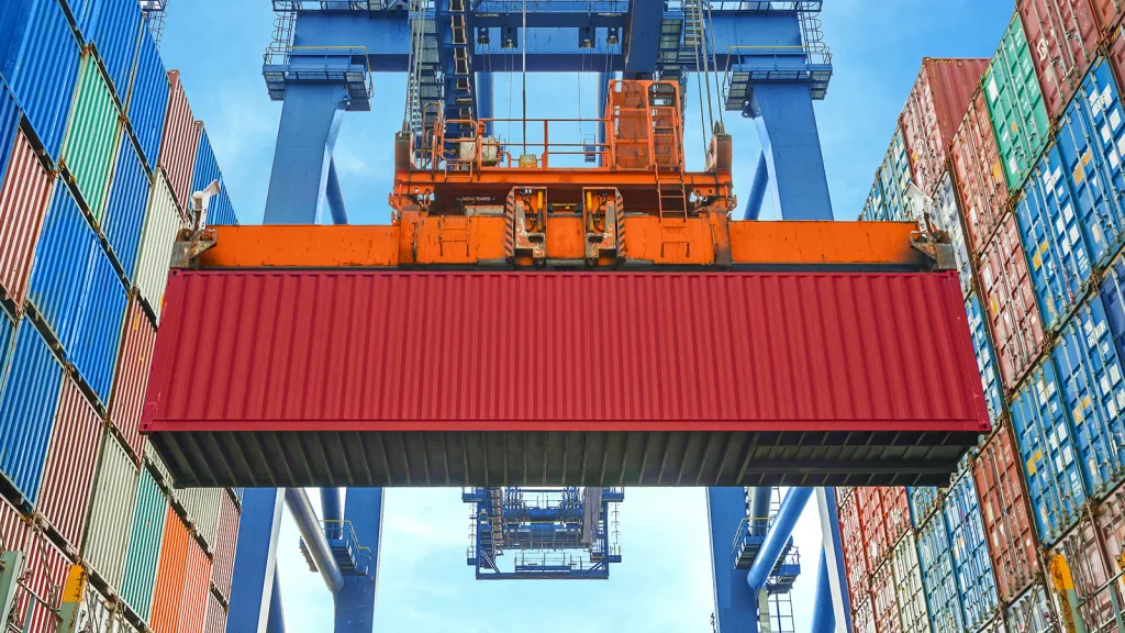 Container wird von einem großen Hafenkran zwischen gestapelten Containern gehoben