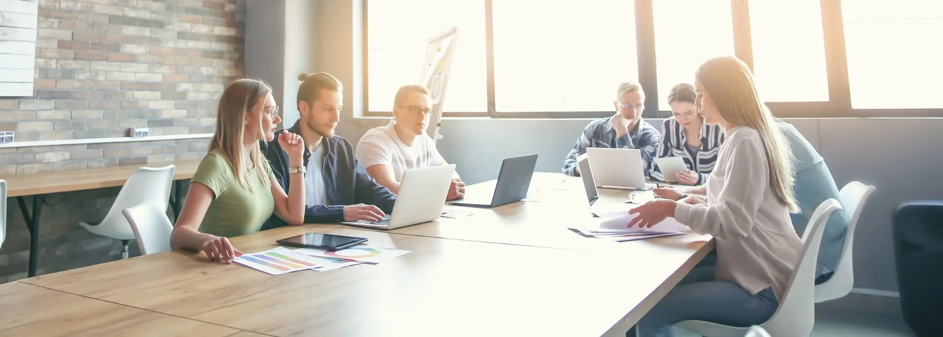 Gruppe von sechs Personen sitzt an einem langen Holztisch in einem modernen, hellen Büro mit großen Fenstern und Ziegelwand und arbeitet gemeinsam an Laptops und Dokumenten